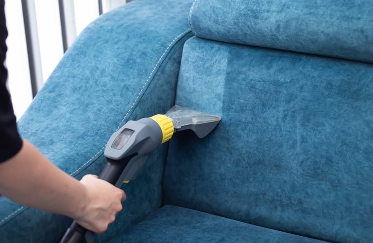 A person is using a vacuum cleaner to clean a blue sofa in a well-lit living room
