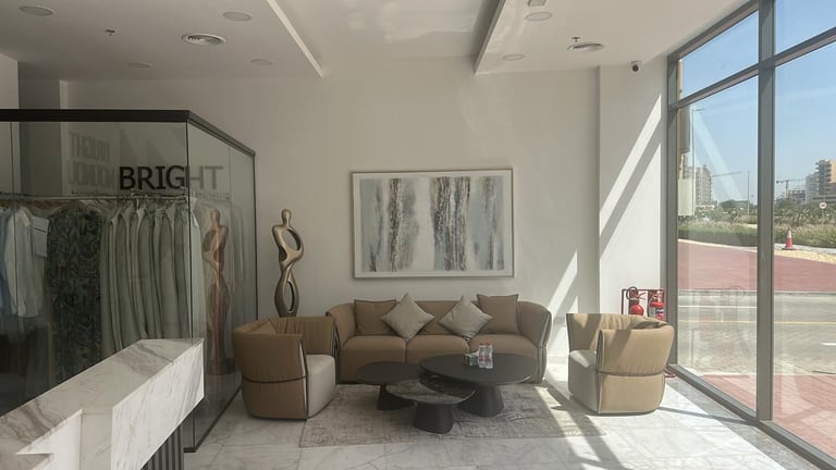 Modern showroom with tan curved sofa, accent chairs, clothing display rack labeled BRIGHT, framed artwork, and floor-to-ceiling windows overlooking cityscape